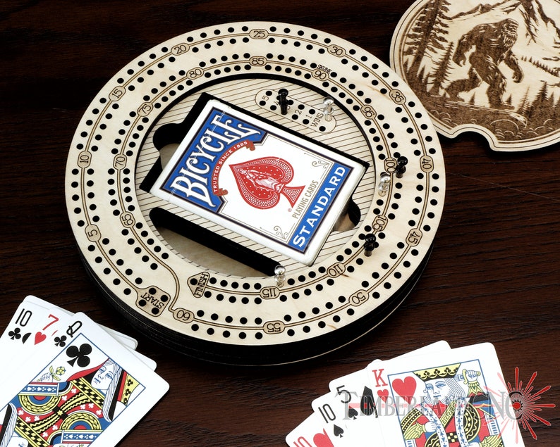 Puede incluir: Un tablero de cribbage de madera con una pista circular y una baraja de cartas Bicycle. El tablero tiene un dise&ntilde;o en blanco y negro con n&uacute;meros y agujeros para clavijas. La baraja de cartas est&aacute; en una caja negra con el logotipo de Bicycle y las palabras "Trusted Since 1885" y "Playing Cards Standard".
