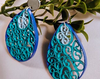 May include: Teardrop-shaped earrings with intricate paper filigree designs. The earrings feature a vibrant color palette of teal, light blue, and dark blue. The filigree is accented with a silver-colored stud. The earrings are displayed against a backdrop of green foliage.