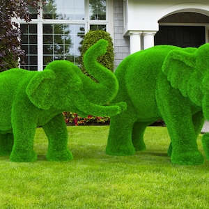 May include: Two green, grass-covered elephant sculptures stand on a green lawn. The larger elephant is standing with its trunk raised, while the smaller elephant is standing with its trunk lowered.