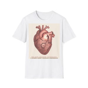 Puede incluir: Camiseta blanca con una ilustración detallada de un corazón humano con elementos mecánicos y una pequeña figura. El texto dice: "THE MOST ADVANCED TECHNOLOGY A HUMAN HEART CHOOSING COMPASSION."