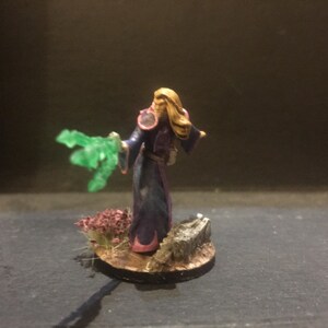Puede incluir: Una figura miniatura pintada de una maga lanzando un hechizo. Lleva una túnica morada y azul con detalles dorados y tiene el pelo largo y rubio. Sostiene un orbe verde brillante en su mano derecha. La miniatura está colocada sobre una base circular con detalles pintados en marrón y verde.