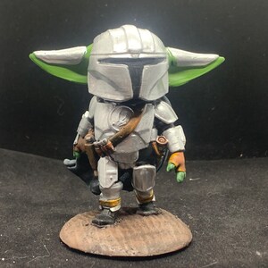 May include: A small, green-skinned figure wearing silver armor and a helmet. The figure is holding a weapon in its right hand and has a cape draped over its back. The figure is standing on a brown base.