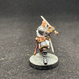 Puede incluir: Una figura miniatura pintada de un guerrero que lleva una armadura blanca y gris, sosteniendo una espada en su mano derecha. La figura está de pie sobre una base circular gris y blanca.