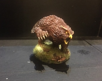 Pode incluir: Uma miniatura pintada de marrom e amarelo que representa uma criatura semelhante a um urso que rosna com grandes olhos de coruja. A miniatura está em pé sobre uma base verde.