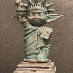 Op de afbeelding: Een groen en gouden standbeeld van het Vrijheidsbeeld, maar met het hoofd en de oren van een Yoda-personage. Het standbeeld houdt een fakkel in de ene hand en een boek met de tekst "Give Me Your Tired" in de andere. Het standbeeld staat op een grijze basis.