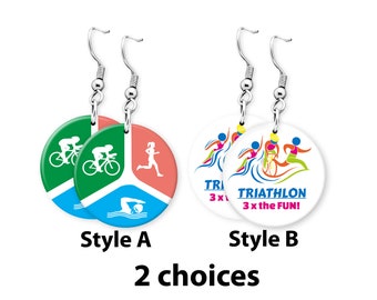 Puede incluir: Dos pares de pendientes con diseños redondos y coloridos. Un par presenta un diseño verde, azul y rosa con un nadador, un ciclista y un corredor. El otro par presenta un diseño blanco con un triatleta y el texto "TRIATHLON 3 x the FUN!"