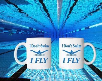 Puede incluir: Dos tazas de café blancas con texto azul y un gráfico de nadador azul. El texto en las tazas dice "I Don't Swim I FLY". Las tazas están sobre una superficie de azulejos azules.