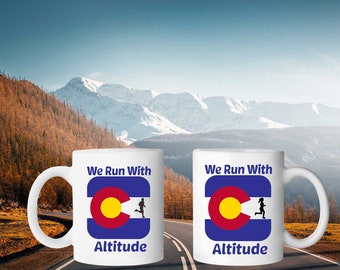 Puede incluir: Dos tazas de cerámica blanca con diseños de la bandera del estado de Colorado en azul y amarillo. Las tazas tienen el texto "We Run With Altitude" impreso en ellas.