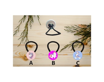 Puede incluir: Tres gomas para el pelo con diseños de unicornio. Las gomas son negras con un botón redondo que presenta un unicornio sobre un fondo rosa, magenta o azul. Las gomas están etiquetadas con A, B y C.