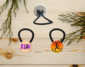 Puede incluir: Dos gomas para el pelo negras con botones redondos de colores. Un botón tiene la palabra "RUN" en morado con una silueta de un corredor. El otro botón tiene un degradado de naranja y amarillo con el texto "LOVE RUNNING" y una silueta de un corredor.