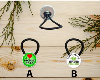 Puede incluir: Dos gomas para el pelo negras con botones redondos verdes y blancos. El botón de la izquierda tiene un corazón rojo y el texto "Me encanta el pickleball". El botón de la derecha tiene el texto "Happy People Play Pickleball".