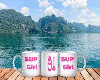Puede incluir: Tres tazas de cerámica blanca con texto rosa y un gráfico de tabla de paddle rosa. Las tazas dicen "SUP Girl". Las tazas están sobre una superficie de madera frente a un lago y montañas.