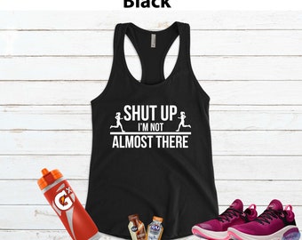 May include: Black tank top with the text "Shut Up I'm Not Almost There" and two running figures. The tank top is on a white wood background with a water bottle, energy gels, and running shoes.