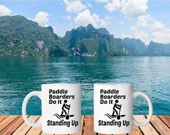 Puede incluir: Dos tazas de cerámica blanca con texto negro y un gráfico de una persona haciendo paddleboard. El texto en las tazas dice "Paddle Boarders Do It Standing Up". Las tazas están sobre una superficie de madera frente a un lago escénico con montañas en el fondo.