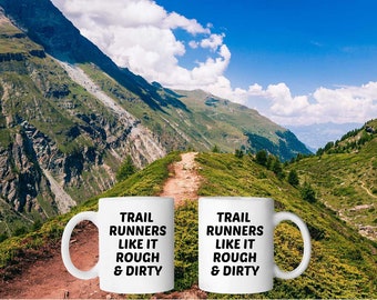 Puede incluir: Dos tazas de café blancas con texto negro que dice "Trail Runners Like It Rough & Dirty". Las tazas están frente a un paisaje montañoso pintoresco con un camino de tierra serpenteando a través de las colinas verdes.