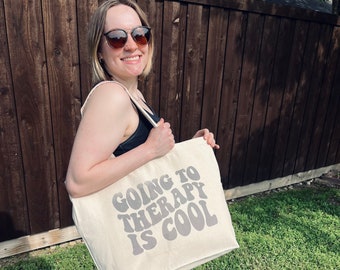 May include: A white canvas tote bag with the text "Going to Therapy is Cool" in a retro font. The text is in gray. The bag is being held by a person wearing sunglasses.