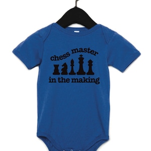 May include: A blue baby onesie with the text "chess master in the making" in black. The text is above a graphic of chess pieces. The onesie is hanging on a black hanger.