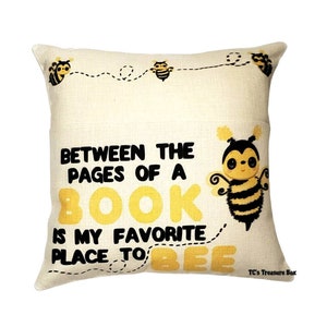 May include: A beige throw pillow with a yellow and black bee illustration and the text "Between the pages of a book is my favorite place to bee".