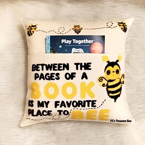 May include: A beige pillow with a pocket for a book. The pillow has a bee design and the text "Between the pages of a book is my favorite place to bee".