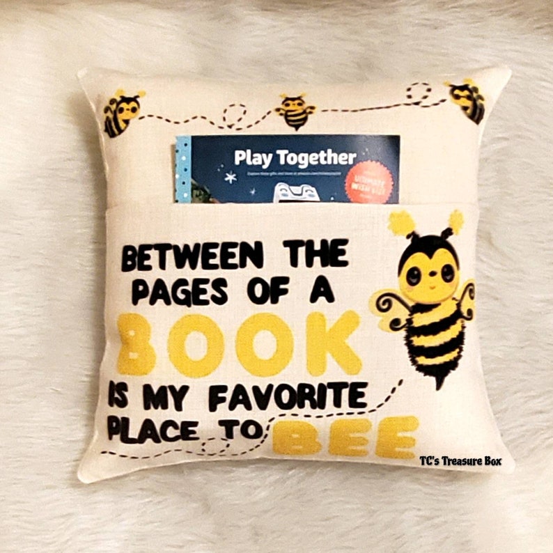 May include: A beige pillow with a pocket for a book. The pillow has a bee design and the text "Between the pages of a book is my favorite place to bee".