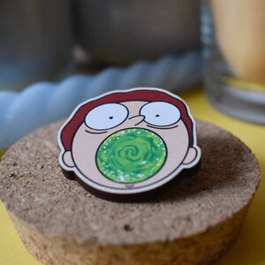 May include: A wooden pin featuring a cartoon character with red hair, wide eyes, and an open mouth. The character is holding a green swirling vortex in their mouth.