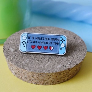 May include: A blue and white enamel pin shaped like a Nintendo Switch console. The pin says "If it makes you happy it's not a waste of time." and has a heart health bar with three full hearts and one half-full heart.