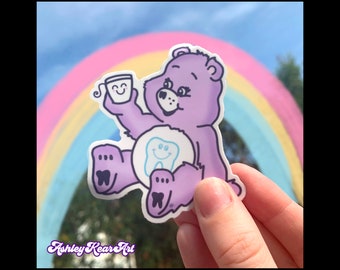 May include: Purple Care Bear sticker with a tooth and a cup with a smiley face. The sticker is in front of a rainbow background.