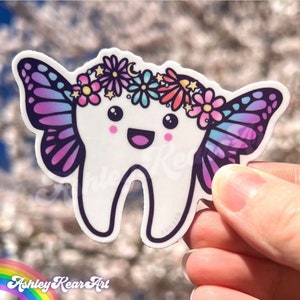 May include: A white tooth with a pink, blue, and purple butterfly on each side. The tooth is wearing a flower crown with pink, blue, and yellow flowers. The tooth has a happy face with big eyes and a smile.