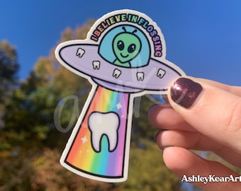 Op de afbeelding: Een sticker met een cartoon-alien in een vliegende schotel met de tekst "I believe in flossing". De schotel is paars en heeft witte tanden rond de rand. Het alien is groen met een grote glimlach. De schotel bevindt zich boven een regenboog-tand met sterren.