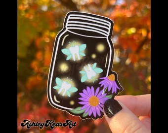 May include: A sticker featuring a black jar with a white rim, containing glowing tooth fairies. The fairies have light blue wings and are surrounded by yellow sparkles. Purple flowers with yellow centers and green leaves are at the bottom. The text "Ashley Rear Art" is at the bottom.