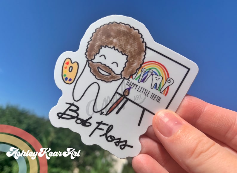 May include: A white sticker with a cartoon drawing of Bob Ross painting a rainbow with three smiling teeth on a canvas. The text on the canvas reads "Happy Little Teeth. Art". The sticker has a black signature that reads "Bob Ross" and the text "Ashley Kear Art" is visible in the background.