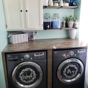 May include: A laundry room with a white cabinet above a wooden countertop. Two black washing machines are on the countertop. The washing machine on the left has a silver door with a wreath and the word "Wash" inside. The washing machine on the right has a silver door with a wreath and the word "Dry" inside.