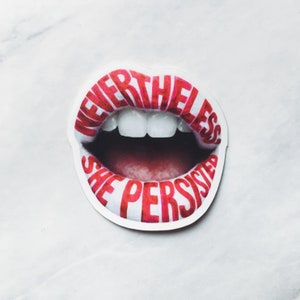 May include: A sticker featuring a stylized image of open lips with the words "NEVERTHELESS SHE PERSISTED" in red lettering. The lips are outlined in white, with the interior of the mouth a deep red. The teeth are white.