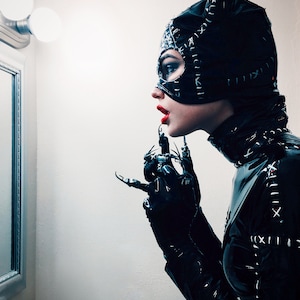 Könnte beinhalten: Eine Person in einem schwarzen Catwoman-Kostüm, einschließlich einer Maske mit spitzen Ohren, einem eng anliegenden Body und Handschuhen mit klauenartigen Verlängerungen. Das Kostüm hat weiße Nähte. Die Person trägt roten Lippenstift.