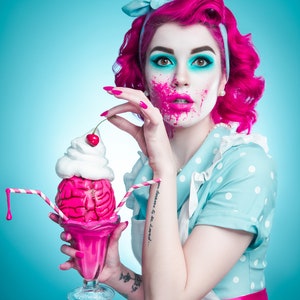 May include: A woman with pink hair and makeup, wearing a blue polka dot dress and a bow, holds a pink milkshake with a brain-shaped ice cream and a cherry on top. The milkshake has striped straws.