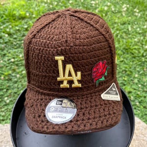 May include: A brown crocheted baseball cap featuring a gold embroidered LA logo on the front and a red rose patch. The cap includes a circular 9FIFTY snapback sticker on the brim and a small white tag that reads Curved Visor Pre-Curved.