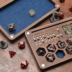 Puede incluir: Una bandeja y organizador de dados de madera con interior de fieltro azul. El organizador tiene ranuras para dados y miniaturas, con el texto "spell slots" visible. Varios dados en colores rojo, negro y transparente están dispersos.