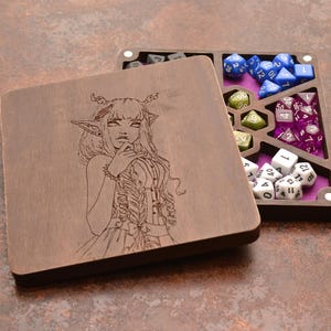 Può includere: Una scatola quadrata in legno per dadi con coperchio rimovibile caratterizzato da un'illustrazione incisa al laser di un personaggio elfico. La base aperta contiene diversi scomparti foderati in feltro viola, contenenti dadi da gioco.