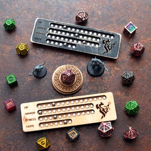 May include: A top-down view showcases a collection of gaming accessories. Two wooden HP, Armor, Stress, and Hope trackers with metal beads are displayed alongside various polyhedral dice in green, yellow, red, and black. Miniature figurines and a wooden dice holder are also present.