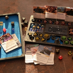 Op de afbeelding: Een tabletop-gameset met dobbelstenen, kaarten en organizers. De set bevat een met blauw vilt gevoerde lade, een dobbelsteenhouder met diverse compartimenten en karakterkaarten. Dobbelstenen zijn in diverse kleuren en vormen.