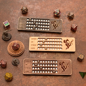 May include: Three wooden RPG trackers with engraved designs and text labels for Armor, HP, Stress, and Hope. Each tracker has small metal ball bearings. Various polyhedral dice and miniature figurines are scattered around the trackers on a brown surface.