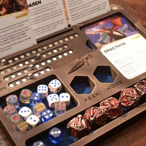 Könnte beinhalten: Ein hölzerner Tisch-Gaming-Organizer mit Würfeln, Karten und Metallkugelmarkern. Der Organizer hat Fächer für Würfel, darunter blaue und weiße Würfel sowie rote Metallwürfel. Karten mit Text sind sichtbar.