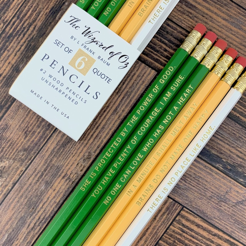 May include: A set of six #2 wood pencils with quotes from The Wizard of Oz. The pencils are green, yellow, and white with gold lettering and red erasers. The pencil box is white with black text.