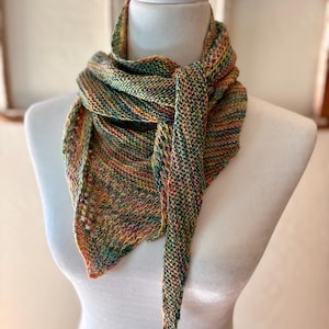 May include: A multicolored knitted scarf displayed on a white mannequin. The scarf features a blend of green, yellow, orange, and red hues. The triangular scarf has a textured knit pattern and a long, pointed end that hangs down.