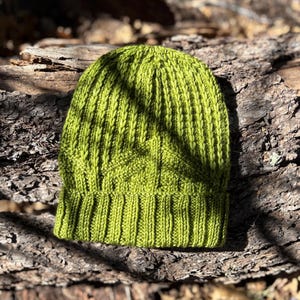 May include: A lime green knit beanie hat with a ribbed design. The hat is made of a soft, textured yarn and is shown on a natural wood surface. The hat is a solid color and has a folded brim.