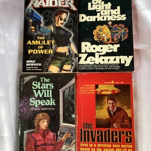 May include: Four paperback books with colorful covers. The first book is titled "Tomb Raider: The Amulet of Power" and features a woman holding a gun. The second book is titled "Creatures of Light and Darkness" and features a red and black illustration. The third book is titled "The Stars Will Speak" and features a woman in front of a computer screen. The fourth book is titled "The Invaders" and features a man running away from a flying saucer.