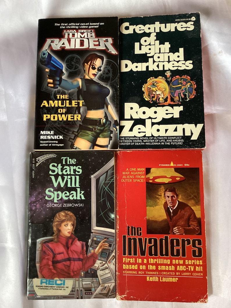 May include: Four paperback books with colorful covers. The first book is titled "Tomb Raider: The Amulet of Power" and features a woman holding a gun. The second book is titled "Creatures of Light and Darkness" and features a red and black illustration. The third book is titled "The Stars Will Speak" and features a woman in front of a computer screen. The fourth book is titled "The Invaders" and features a man running away from a flying saucer.