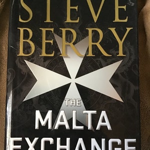 May include: A black book cover with a gold title that reads "Steve Berry" and a white Maltese cross. The title of the book is "The Malta Exchange" and it is a novel.