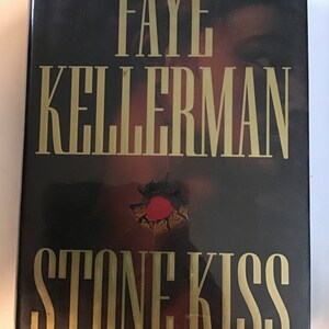 May include: A hardcover book with a black cover and gold lettering. The title is "Stone Kiss" by Faye Kellerman. The cover features a close-up of a person's face with a bullet hole in the forehead.