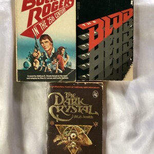 May include: Three paperback books, one with a red and black cover featuring the text "The Blood", one with a red and white cover featuring the text "Buck Rogers in the 25th Century", and one with a brown cover featuring the text "The Dark Crystal" and a graphic of a creature with a large eye.